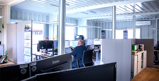 Modernes Großraumbüro in einer Logistikspedition mit mehreren Arbeitsplätzen, Trennwänden und großen Fenstern. Mitarbeitende sitzen an Schreibtischen vor mehreren Monitoren und arbeiten am Computer. Helle Containerbauweise, Holzfußboden, Aktenordner und Büroausstattung sorgen für eine strukturierte, funktionale Arbeitsumgebung.