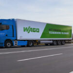 Blauer Sattelzug mit WAGO-Auflieger fährt auf einer mehrspurigen deutschen Autobahn, aufgenommen seitlich in Bewegung mit Leitplanke im Vordergrund und weiter Landschaft im Hintergrund.