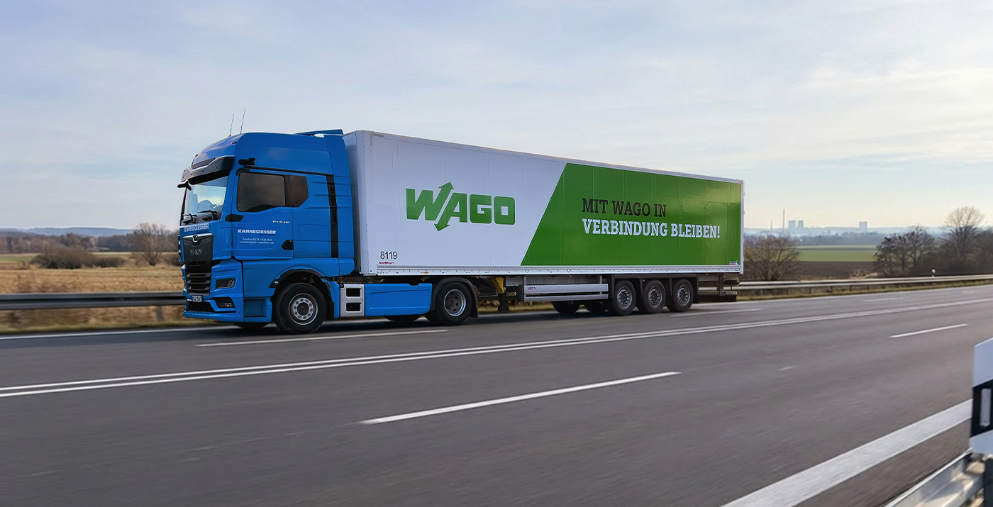 Blauer Sattelzug mit WAGO-Auflieger fährt auf einer mehrspurigen deutschen Autobahn, aufgenommen seitlich in Bewegung mit Leitplanke im Vordergrund und weiter Landschaft im Hintergrund.