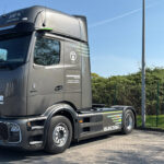 Grauer Mercedes E-LKW ohne Auflieger. Stehen auf dem Betriebsgelände. Im Hintergrund Firmenfahnen zu sehen