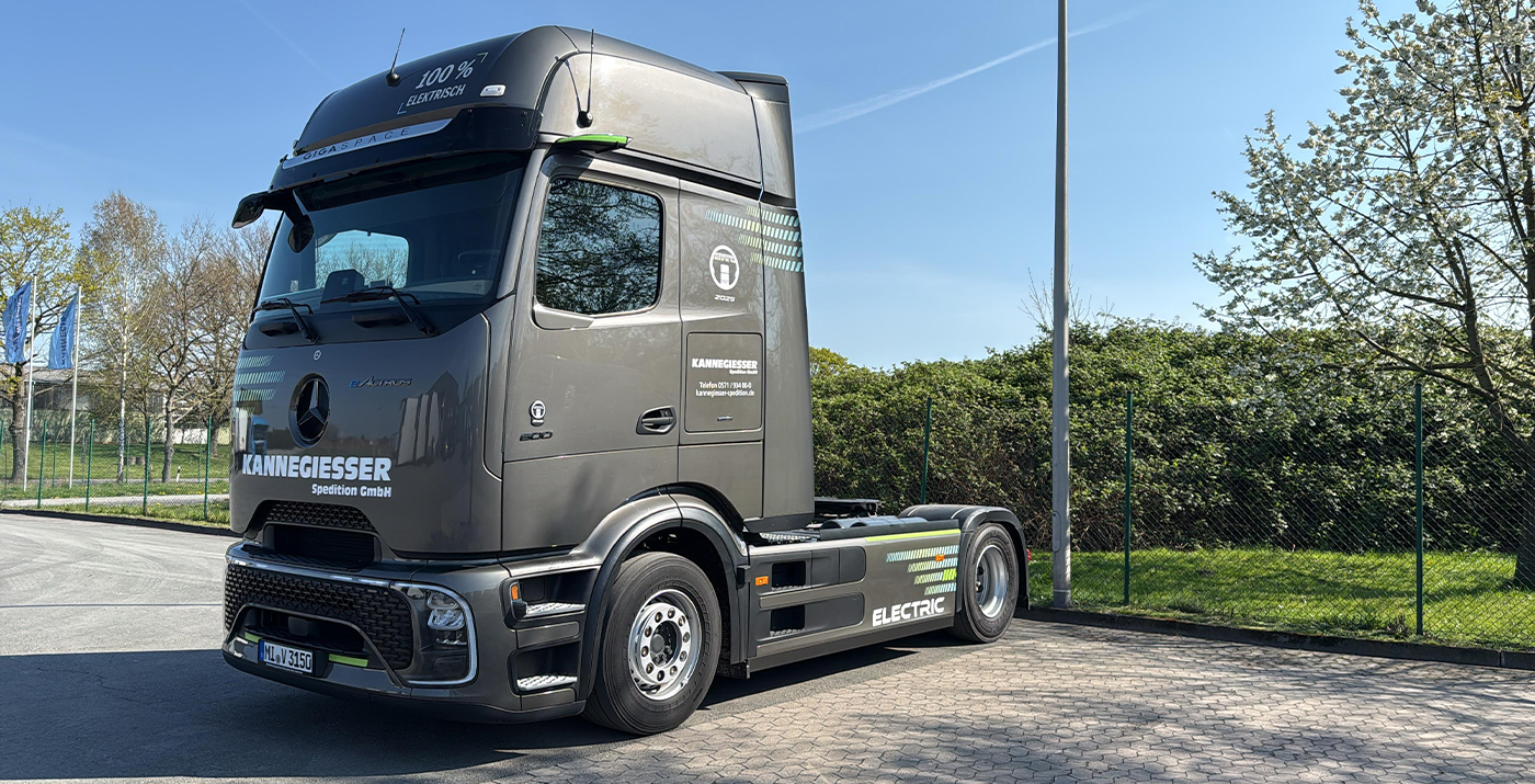 Grauer Mercedes E-LKW ohne Auflieger. Stehen auf dem Betriebsgelände. Im Hintergrund Firmenfahnen zu sehen